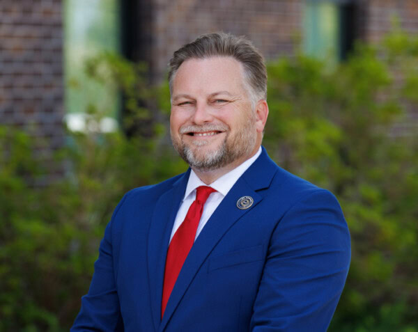 Smiling man in a blue suit and red tie stands outside by greenery and a brick building in St. George, LA.
