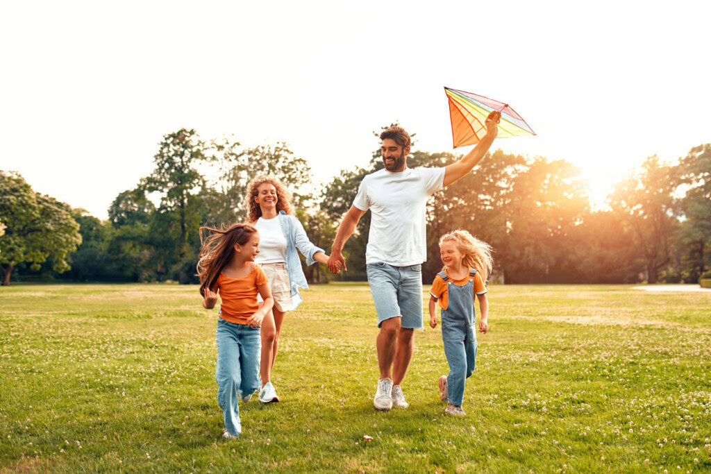 A family of four runs on grass in a St. George park as the father holds a colorful kite aloft.
