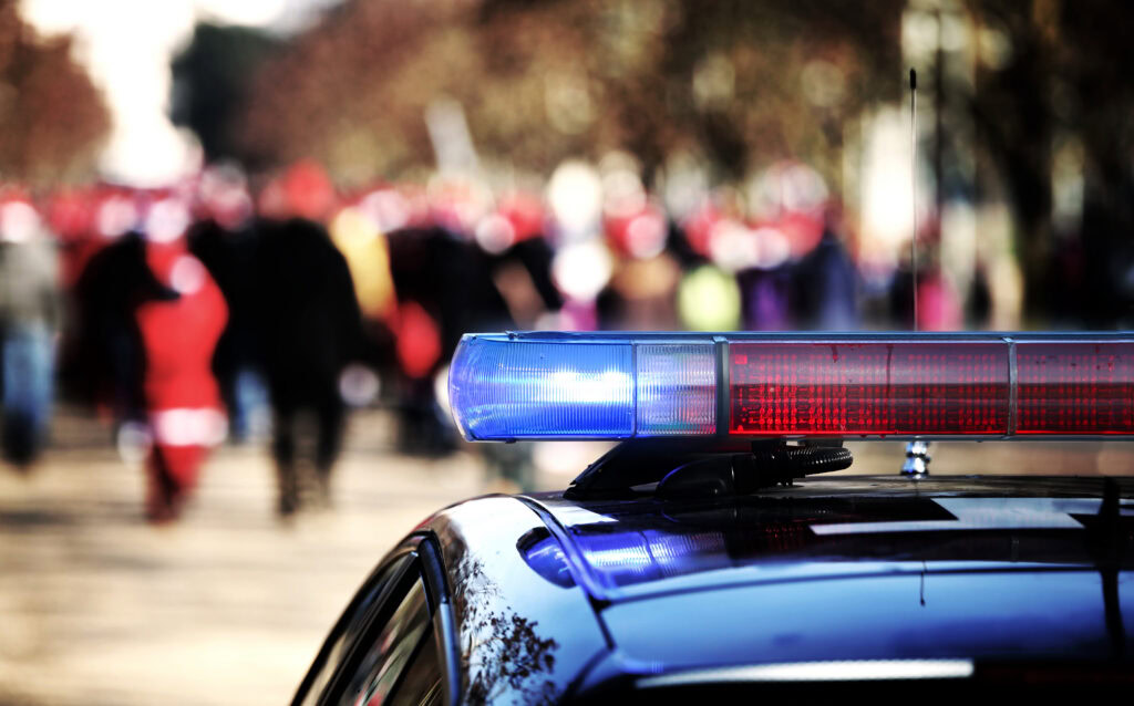 A police car with blue and red lights is parked on a St. George street, crowd blurred behind.