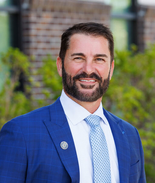 Man with dark hair and beard in a blue suit stands outside in St. George LA by brick wall and greenery.