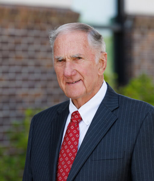 An older man in a dark pinstripe suit and red tie stands outside in St. George, LA, near a blurred building.