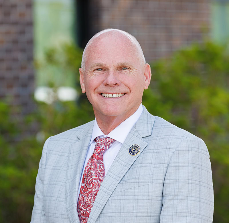 A bald man in a gray plaid suit and red tie smiles outside in St. George LA, with greenery and a brick wall behind him.