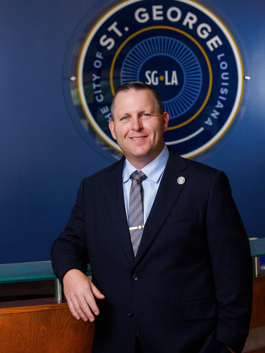 Smiling man in suit stands before a circular City of St. George LA sign.